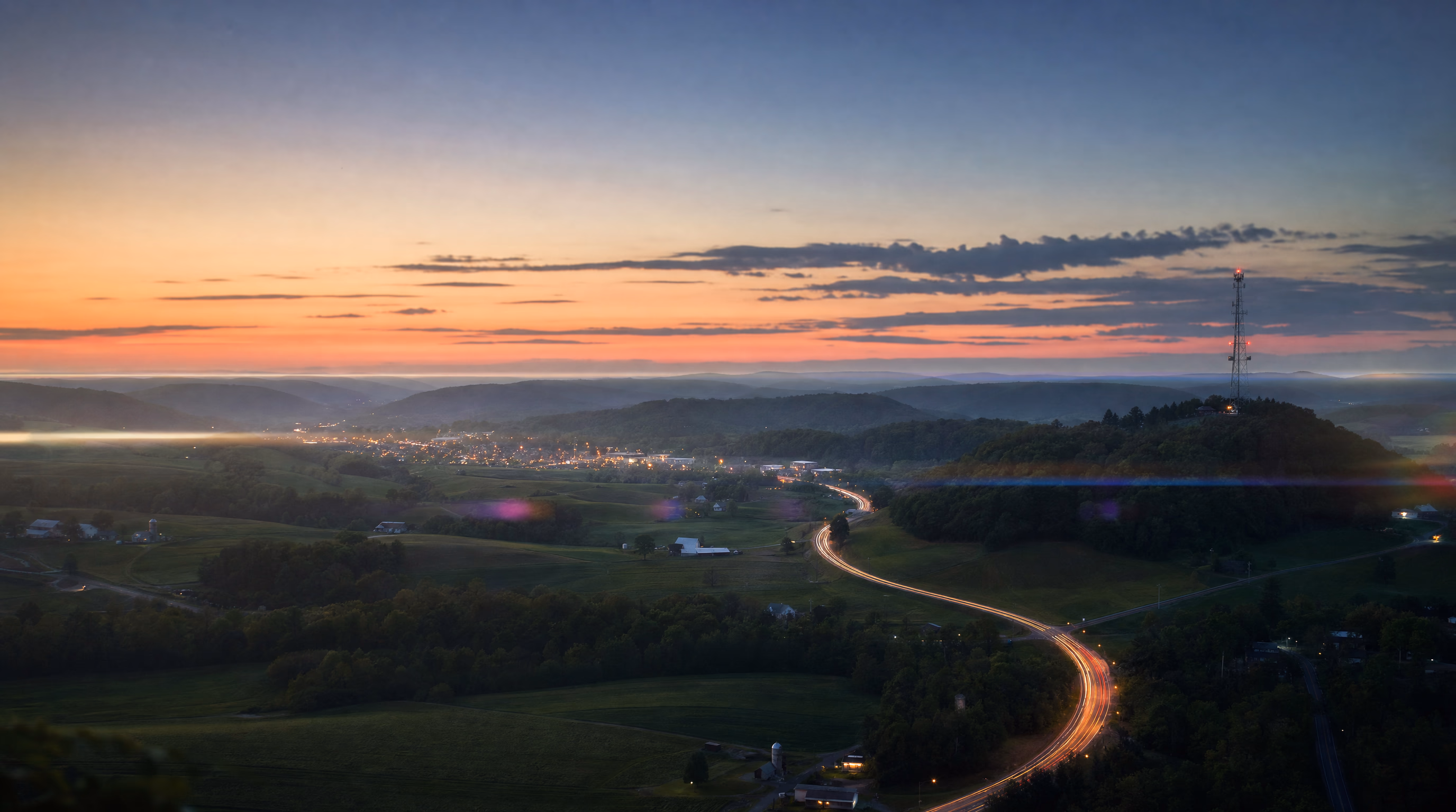 Elevated view over a rural American valley at dusk, a highway winding through farmland toward a glowing town in the distance